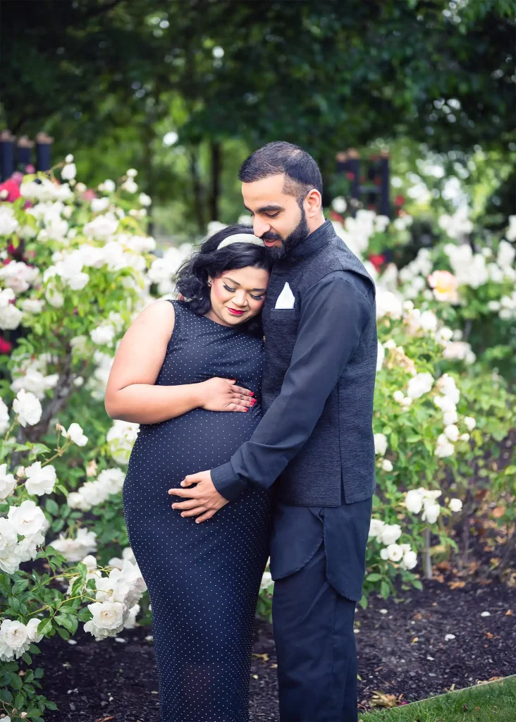 Maternity couple standing together in a garden with white blooms, partner embracing the baby bump