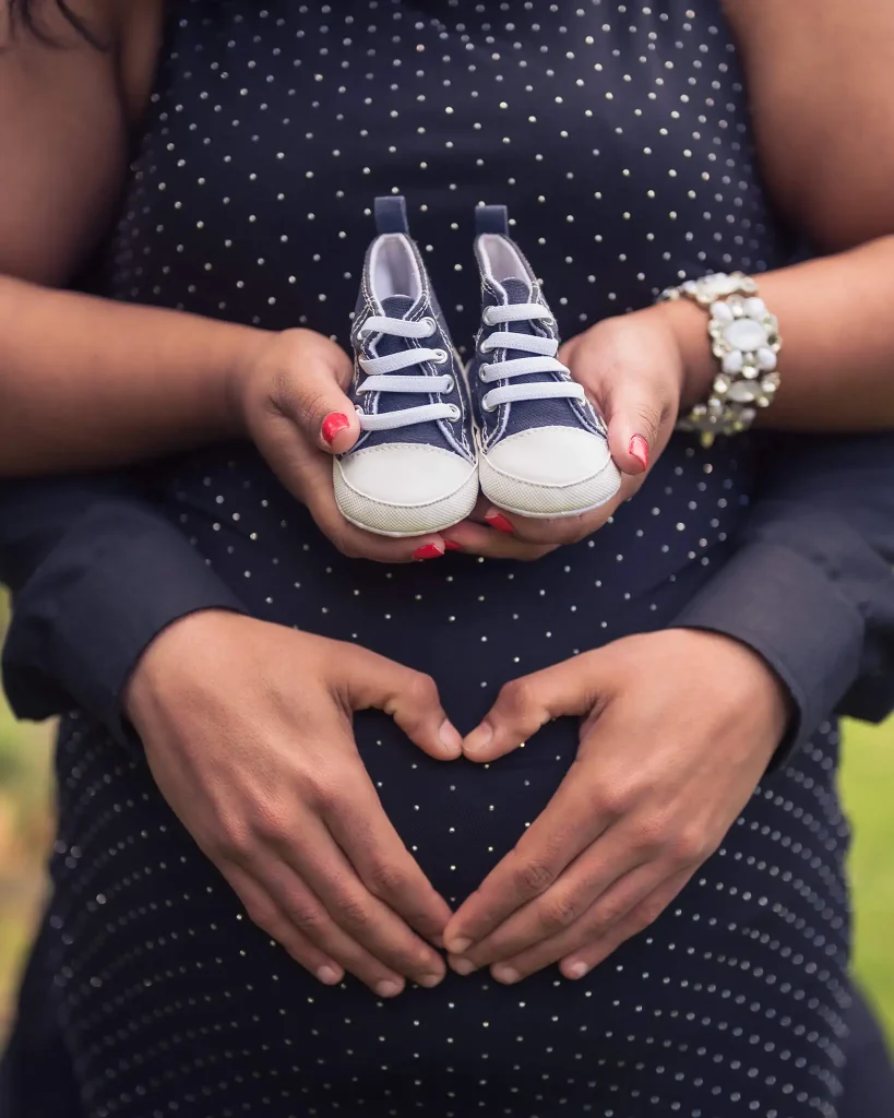 Close-up of pregnant belly with partner’s hands forming a heart and tiny baby shoes held above