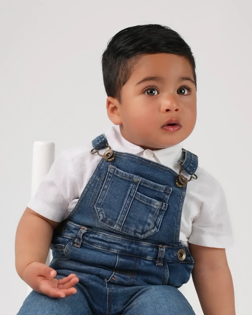 Studio portrait of a toddler boy in denim dungarees sitting on a white chair in Christchurch